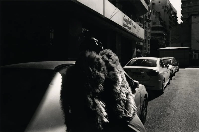 Black and white outdoor shot of an adult wearing a fur coat standing next to a parked car on a city street, with nearby buildings.