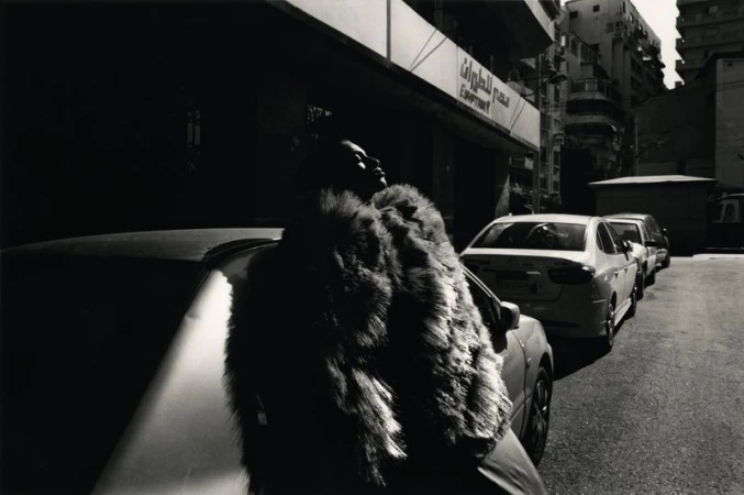 Black and white outdoor shot of an adult wearing a fur coat standing next to a parked car on a city street, with nearby buildings.