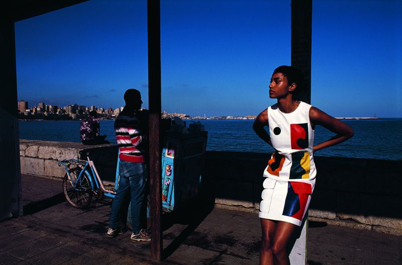 A woman poses in a patterned dress next to the ocean, with a man and bicycle visible in the background along a city waterfront.
