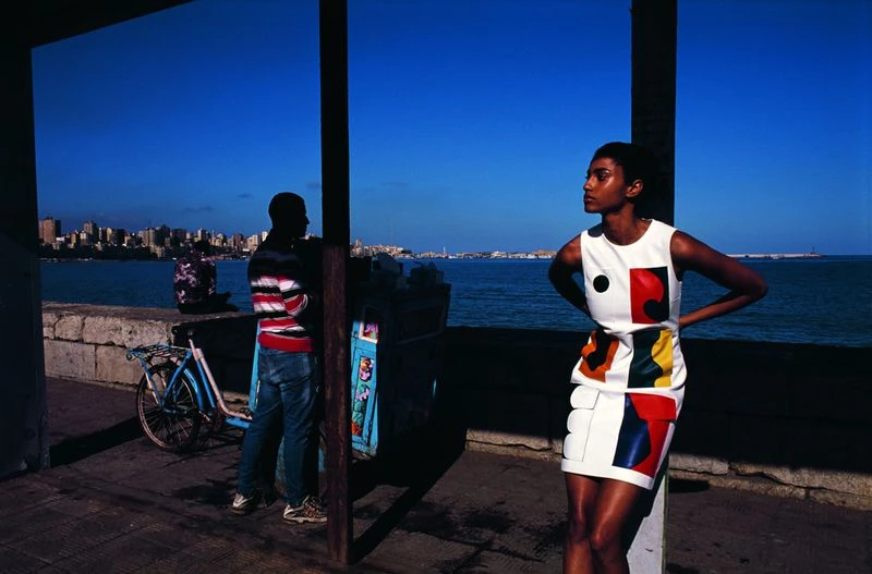 A woman poses in a patterned dress next to the ocean, with a man and bicycle visible in the background along a city waterfront.