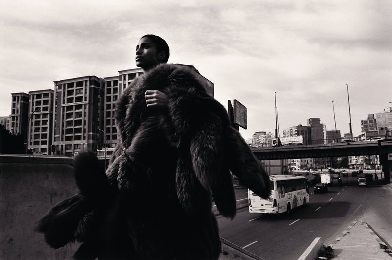 A person in a fur coat stands on a bridge above a road with cars and buildings, photographed in black and white with high contrast.