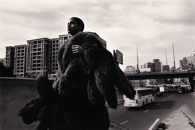 A person in a fur coat stands on a bridge above a road with cars and buildings, photographed in black and white with high contrast.
