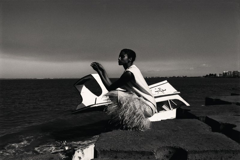 Monochrome portrait of a woman squatting on a stone structure near the ocean with a partially submerged boat in the background.