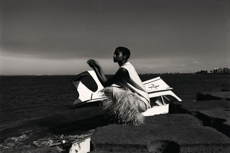 Monochrome portrait of a woman squatting on a stone structure near the ocean with a partially submerged boat in the background.