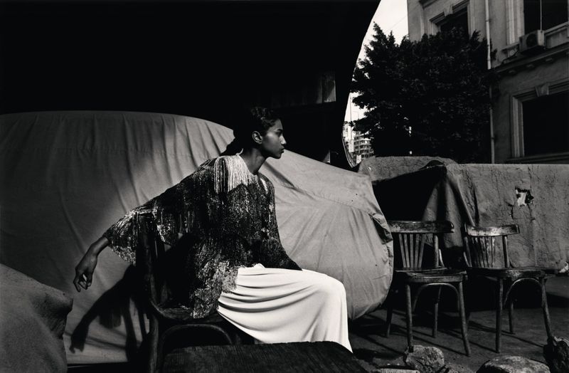 Fashion shot in black and white features a model in an embellished top and wide-leg pants, seated outdoors near chairs and a building.