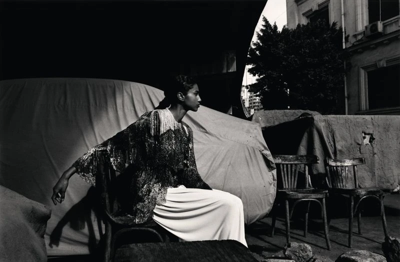 Fashion shot in black and white features a model in an embellished top and wide-leg pants, seated outdoors near chairs and a building.
