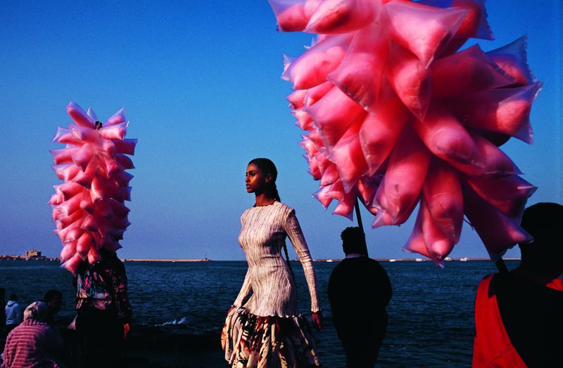A fashion shot features a woman in an elegant dress standing by the ocean, with sellers of pink cotton candy in the background.