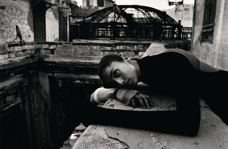 Monochrome portrait of a woman in a dark outfit reclining on a weathered stone structure in a building with an arched dome.