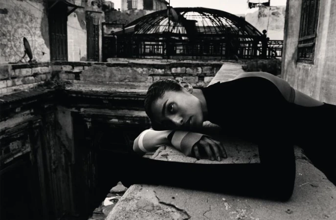 Monochrome portrait of a woman in a dark outfit reclining on a weathered stone structure in a building with an arched dome.