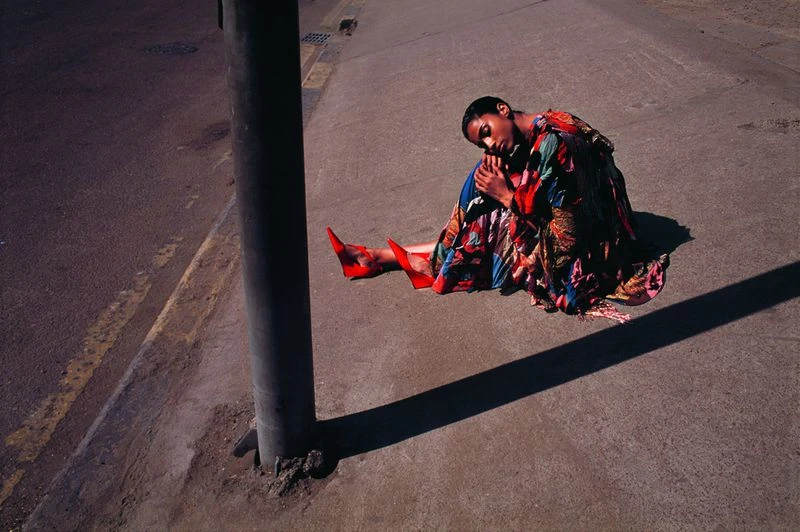 A woman in a colourful dress and red high heels sits on the ground next to a pole, with hard shadows from the sunlight.