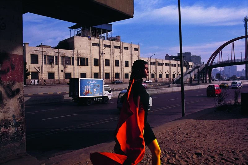 Woman in vivid orange shawl walks in urban environment, showcasing contrasting colors against a cityscape backdrop with a transport truck.