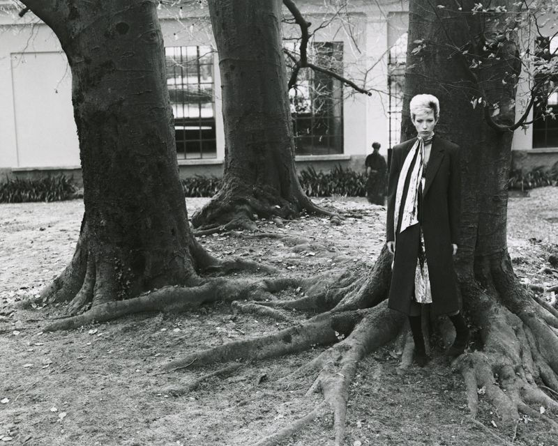 Monochrome portrait of a blonde woman in a black coat and striped scarf standing near large trees with visible roots.