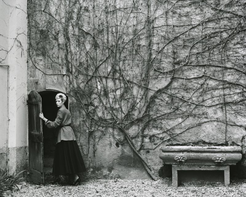Monochrome image captures a woman standing in a doorway with a stone wall covered in vines and a stone planter.