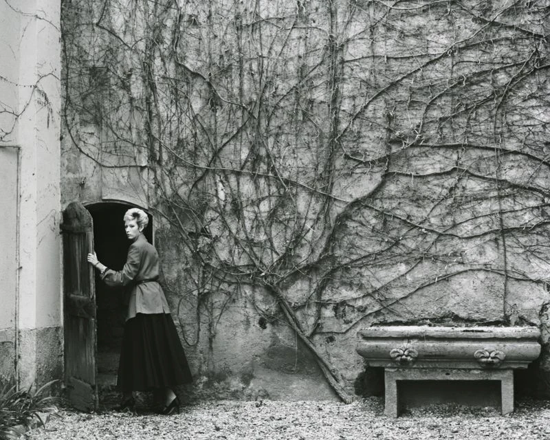 Monochrome image captures a woman standing in a doorway with a stone wall covered in vines and a stone planter.