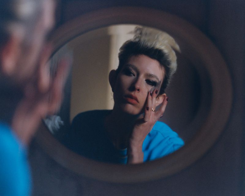 Reflection in oval mirror of adult with fair skin applying dark makeup with fingers indoors in a private residence.