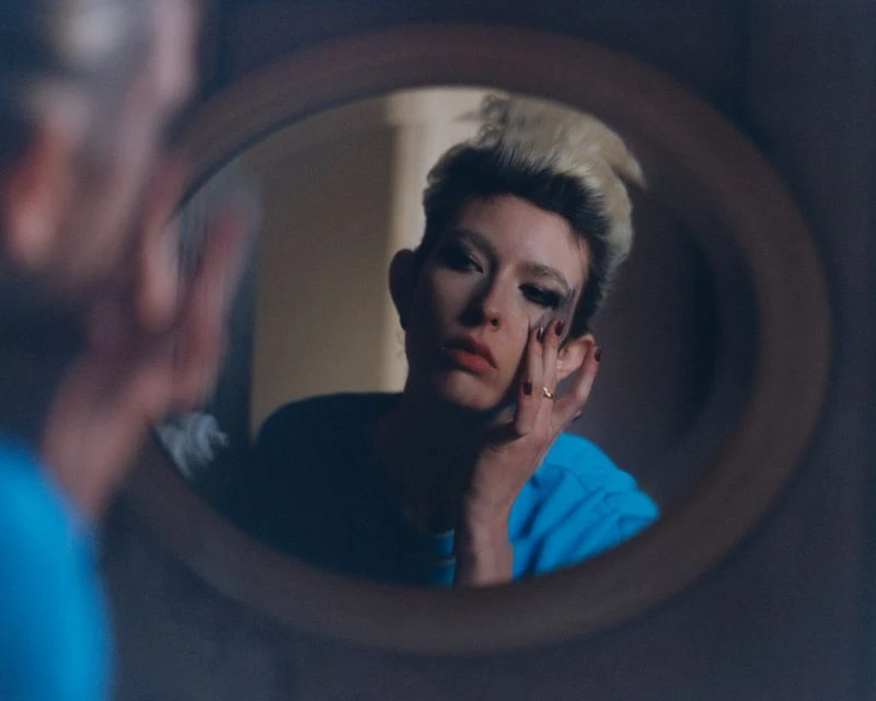 Reflection in oval mirror of adult with fair skin applying dark makeup with fingers indoors in a private residence.