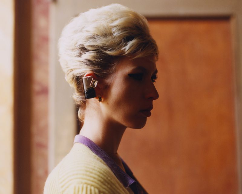 Profile shot of a woman with blonde hair wearing a yellow sweater and large geometric earrings against a blurred, warm-toned background.