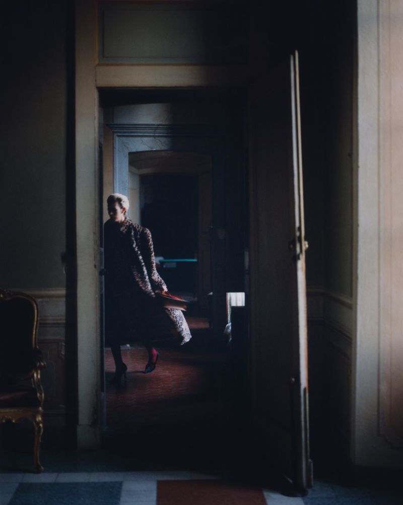 Woman in patterned coat and red tights standing in doorway of interior space with a chair on the left and dark hallway behind her.