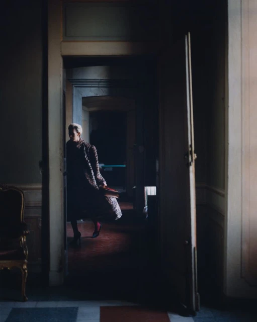 Woman in patterned coat and red tights standing in doorway of interior space with a chair on the left and dark hallway behind her.