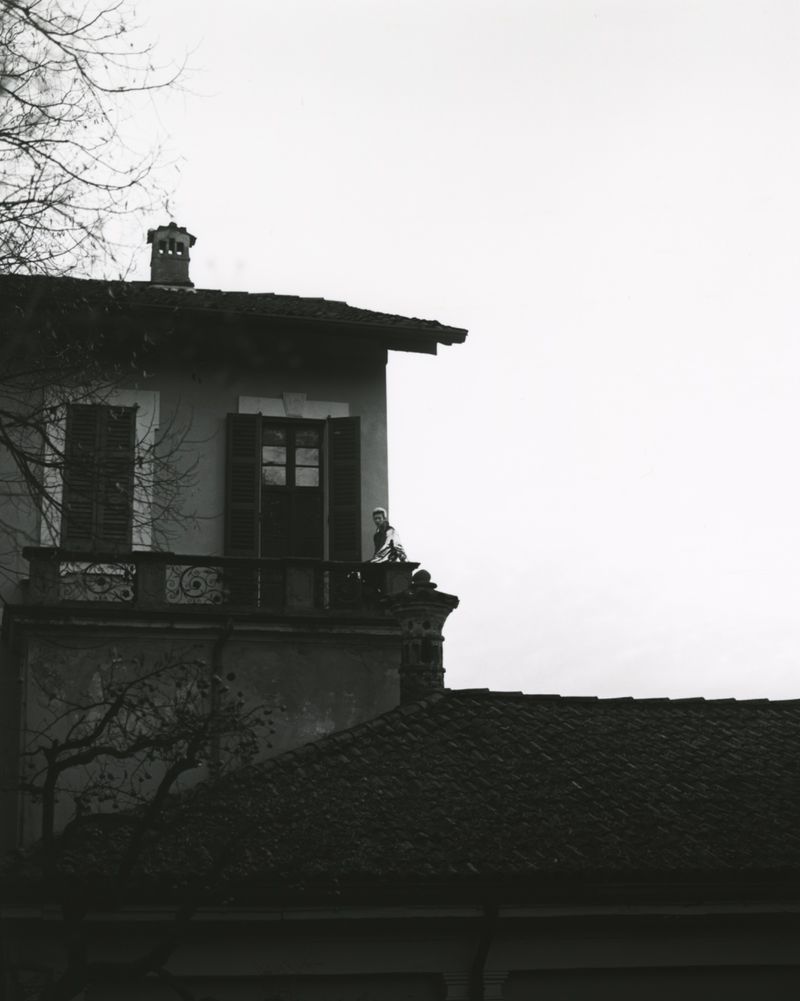 Black and white photo of a person standing on the balcony of a building, with a visible rooftop and bare trees in the background.