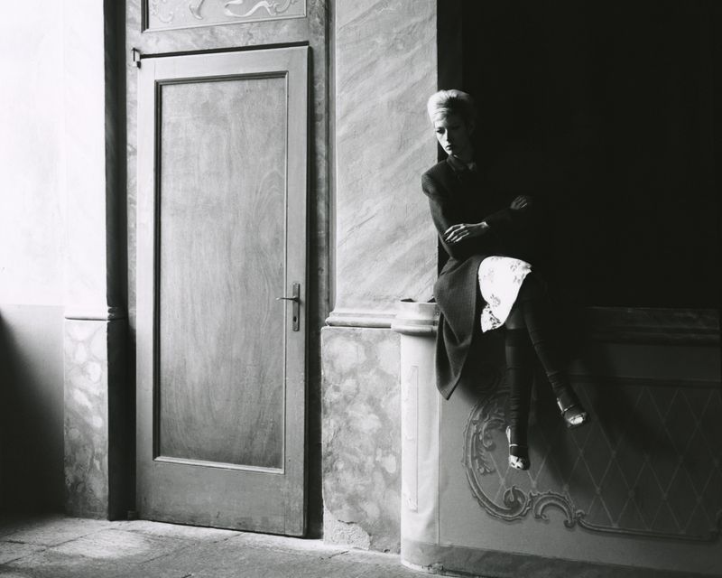 Monochrome photograph of a stylish woman sitting on ledge in a hall, door to left, shot in subdued lighting with elegant wall detailing.