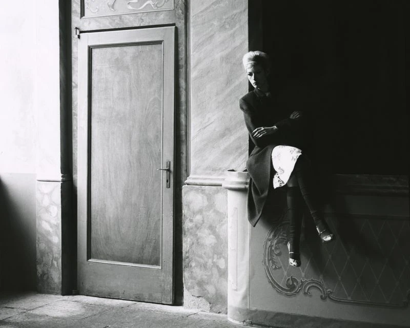 Monochrome photograph of a stylish woman sitting on ledge in a hall, door to left, shot in subdued lighting with elegant wall detailing.