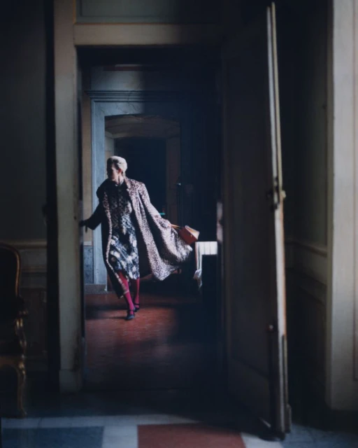 An adult female with short blonde hair and a leopard-print coat is walking through a doorway in a dimly lit interior setting.