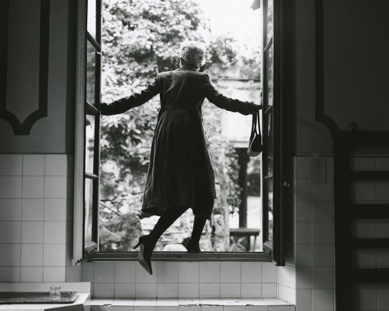 Woman in coat and heels climbing out of a window into a garden on a black and white photo, holding a purse.
