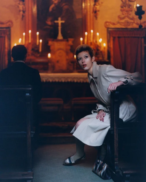 Woman in a beige trench coat sits in a church pew. A man sits in front of her, with candles lit on the altar in the background.