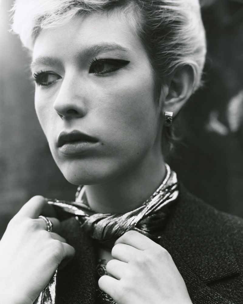 Close-up of a stylish adult female with short blonde hair and distinctive winged eyeliner, wearing a patterned scarf and grey coat.