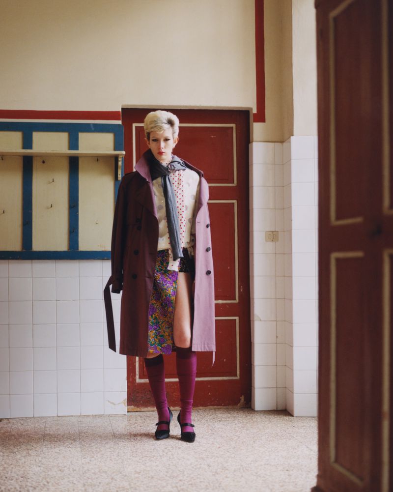 Fashion editorial shot of a blonde model in a colorful outfit featuring a trench coat, mini skirt, stockings, and Mary Jane shoes.