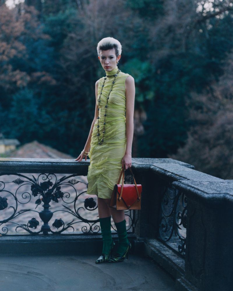 A stylish woman poses outdoors in a bright yellow dress, green stockings, and a red and brown handbag against a dark backdrop.