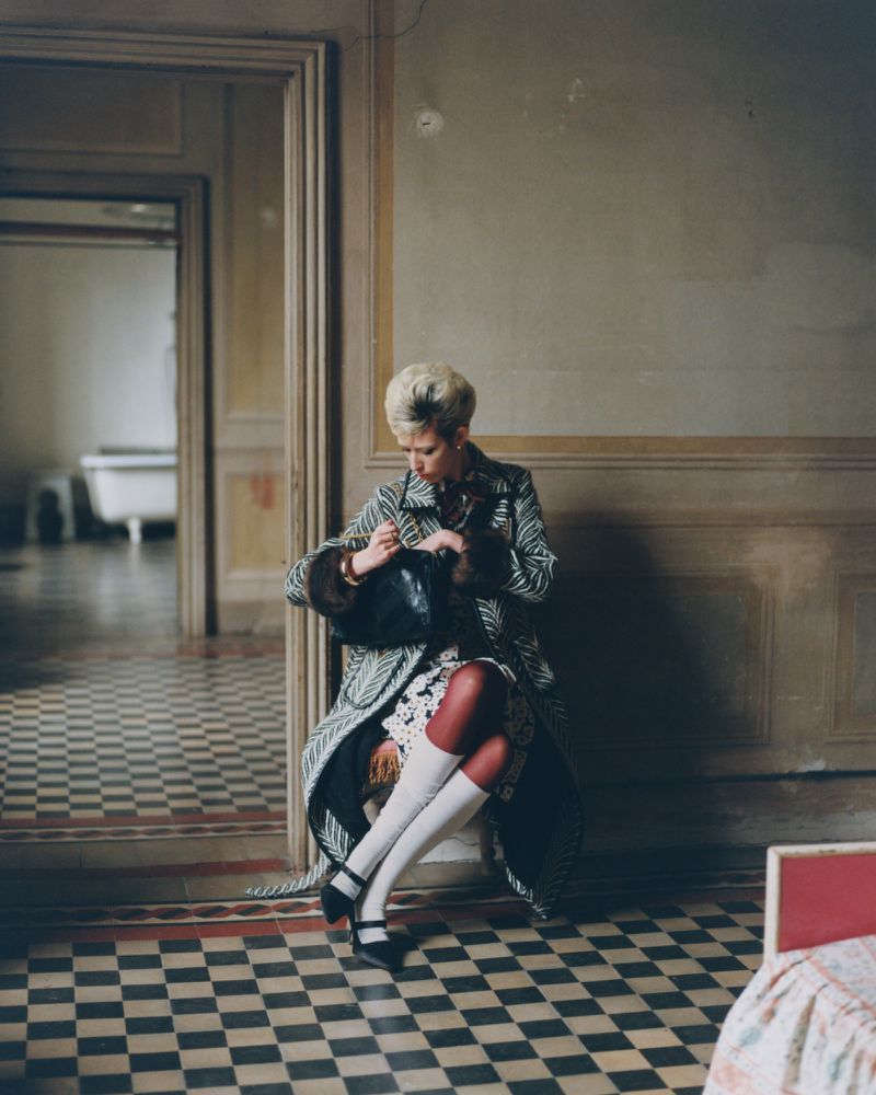 Woman in a patterned coat sits in an empty room with a tiled floor, holding a purse, viewed through a doorway with a glimpse of a bathtub.