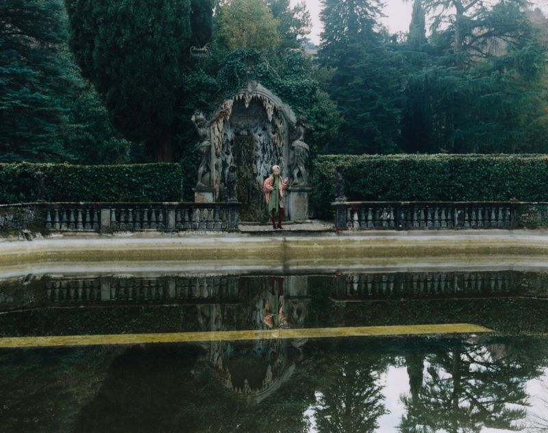 Woman standing near an ornate fountain in a garden setting reflected in a pond, surrounded by lush greenery and trees.