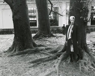 Model standing between large tree trunks with exposed roots, wearing a dark coat and scarf in a park setting.