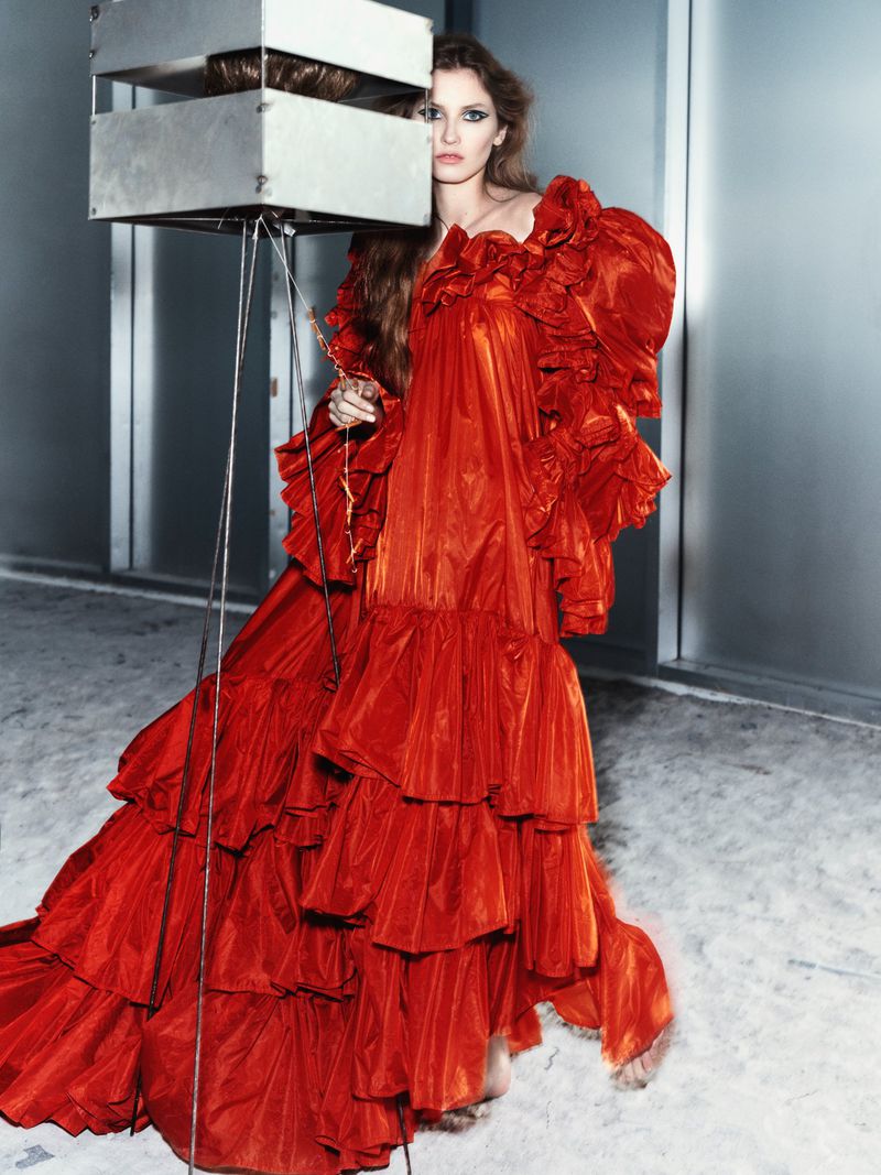 Adult female in a ruffled red gown poses in front of a metal structure in a fashion photography shot against a neutral gray wall.