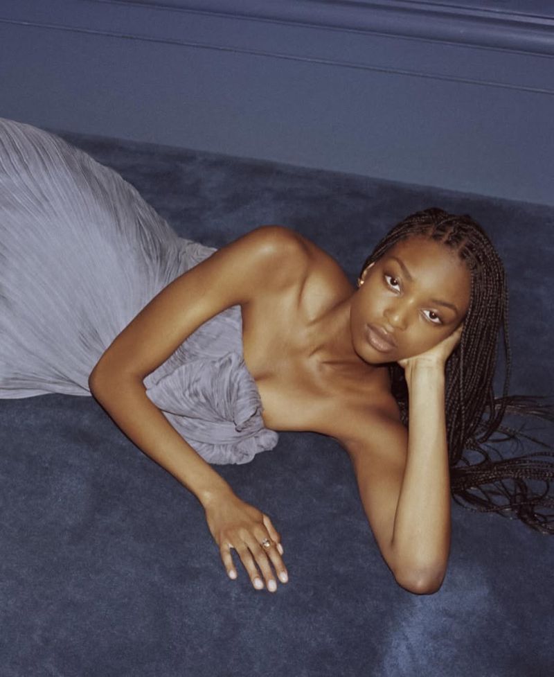 Woman with braids, wearing a grey strapless dress, lies on a bluish-grey surface in a studio, resting her head on her left hand.