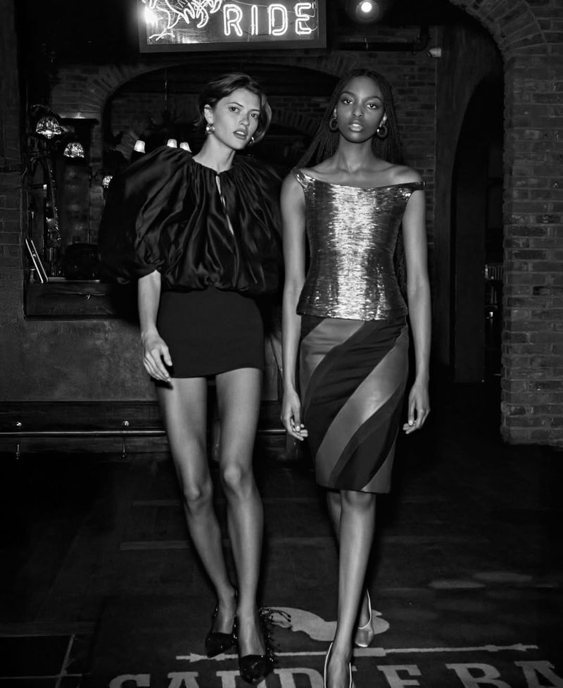 Two fashion models showcase dresses in a dimly lit bar with a brick archway. One wears a dark top and skirt; the other a metallic top and diagonally striped skirt.