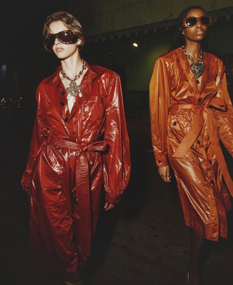 Two fashion models in red and orange trench coats, large sunglasses, and statement necklaces walking in an outdoor environment at night.