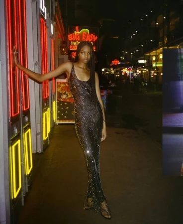 An African American woman with braided hair poses in a shimmering gown at night on a city street with colorful neon signs.