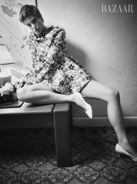 Black and white fashion shot of a woman in a floral dress and white heels, sitting on a table next to a table lamp and flowers.