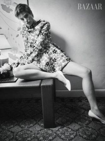 Black and white fashion shot of a woman in a floral dress and white heels, sitting on a table next to a table lamp and flowers.