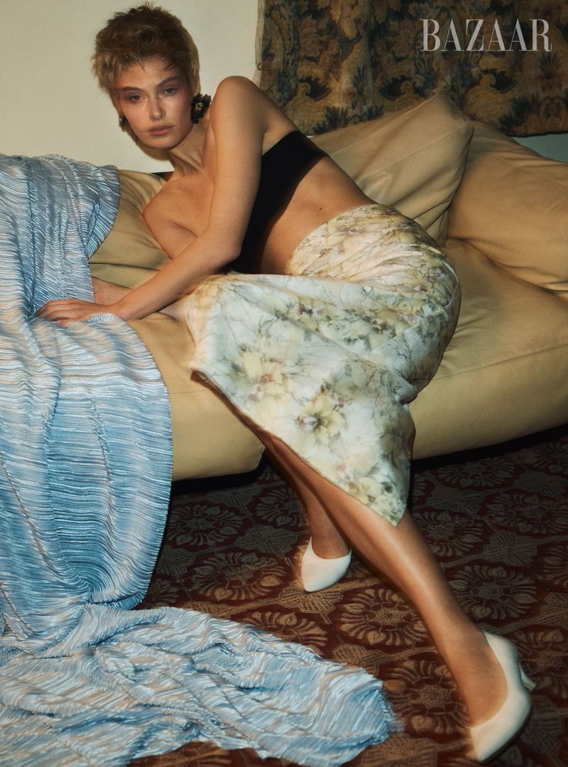A fashionable young woman reclines on a sofa wearing a black top, floral skirt and white heels in a studio shot with a patterned background.