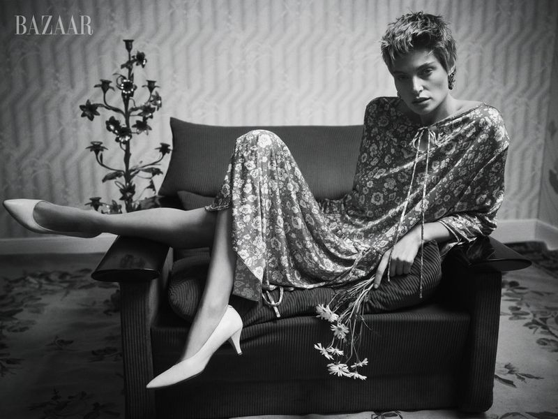 Black and white fashion shot of a woman in a floral dress, seated on a chair with a candelabra and 'BAZAAR' logo visible.