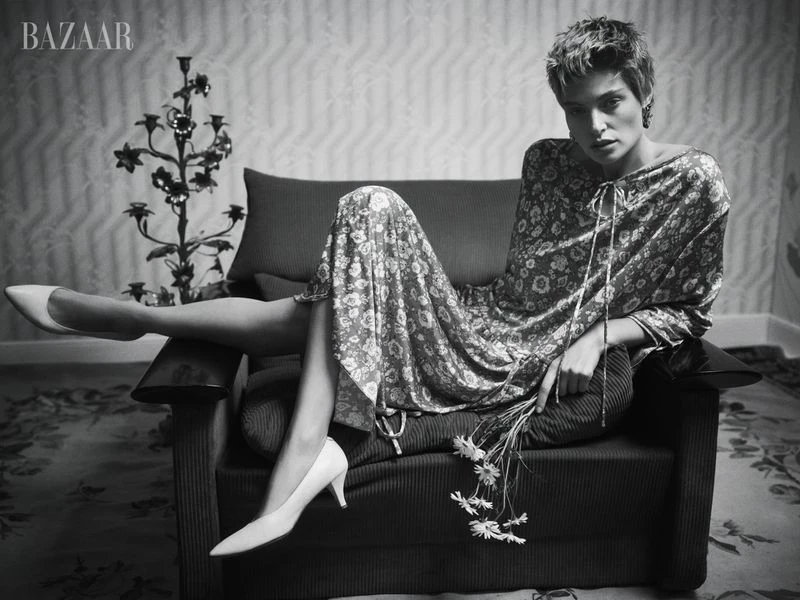 Black and white fashion shot of a woman in a floral dress, seated on a chair with a candelabra and 'BAZAAR' logo visible.