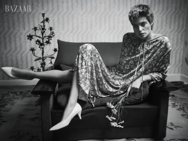 Black and white fashion shot of a woman in a floral dress, seated on a chair with a candelabra and 'BAZAAR' logo visible.