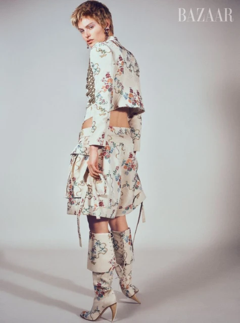 A fashion model in a floral-patterned jacket, skirt, and boots stands confidently in a studio, turning slightly to look over her shoulder.