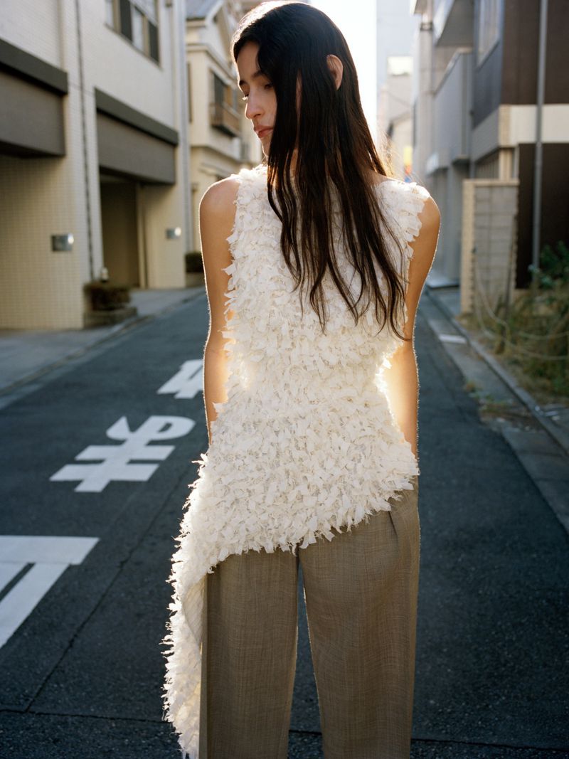 Brunette woman poses in a textured white top and trousers in an outdoor setting on a street with building in the background.