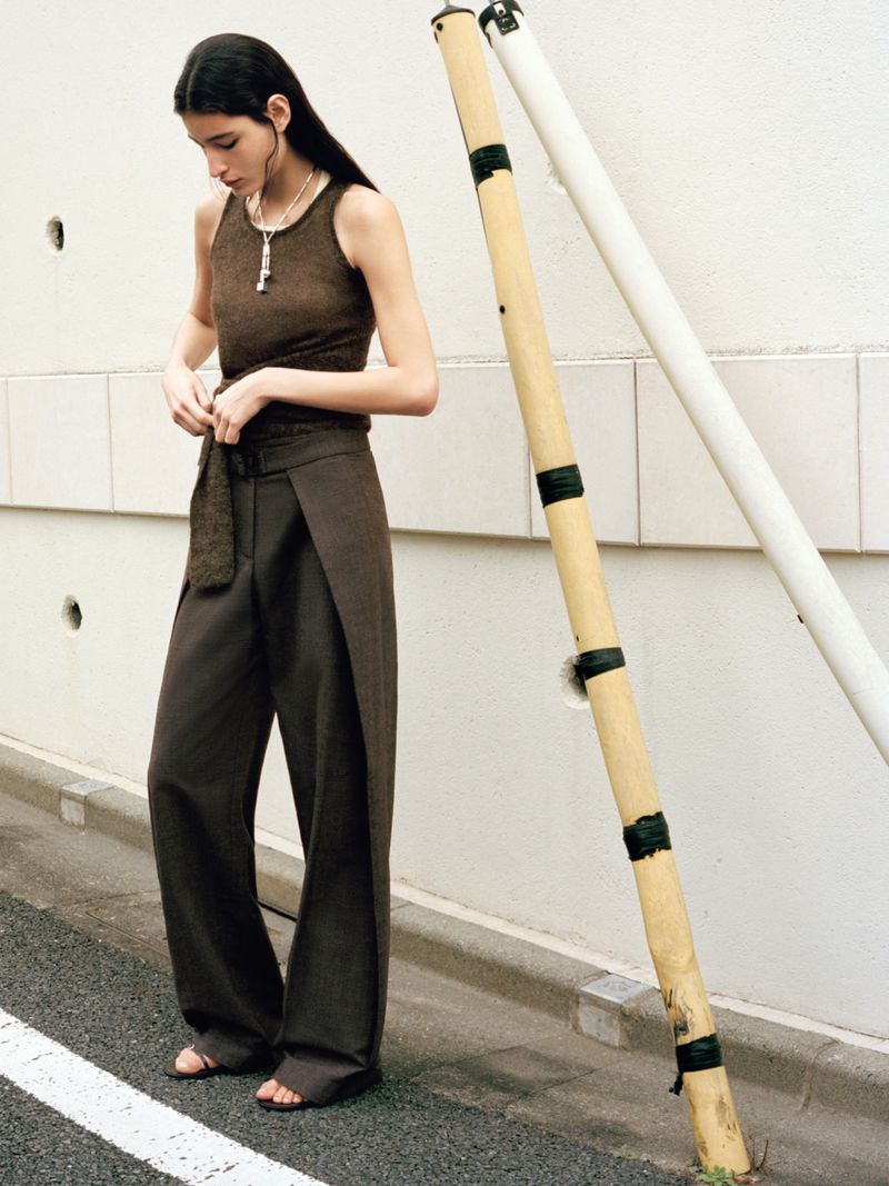 A slender woman in a brown top and dark pants stands outside, the scene is lit with soft light, accentuating the urban setting.