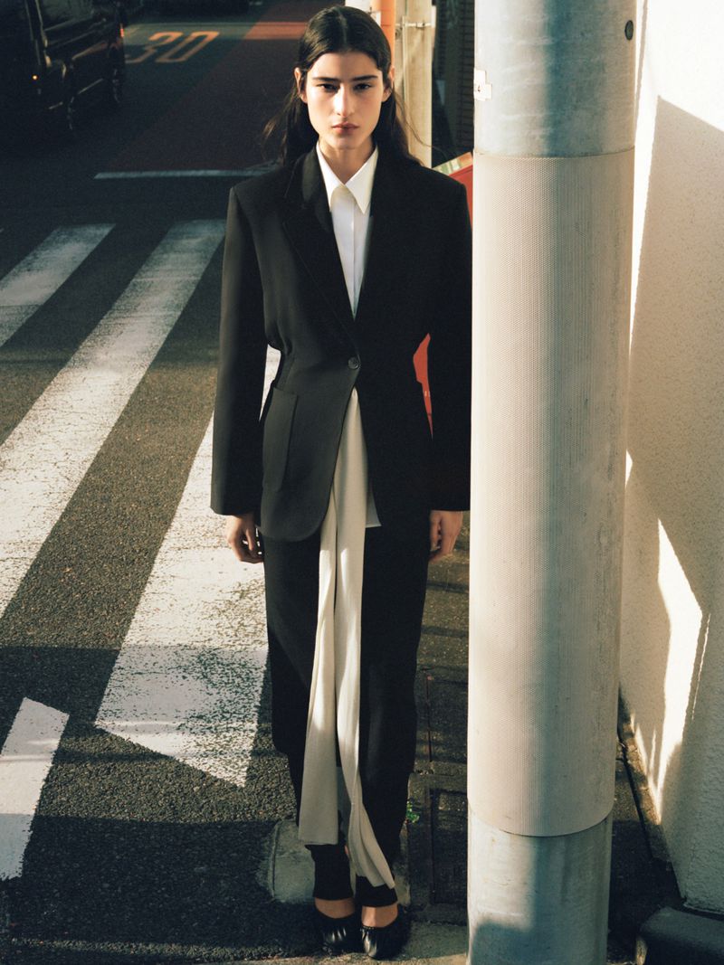 A model in black blazer and pants poses on a crosswalk next to a grey post, showcasing fashion in urban environment with natural light.